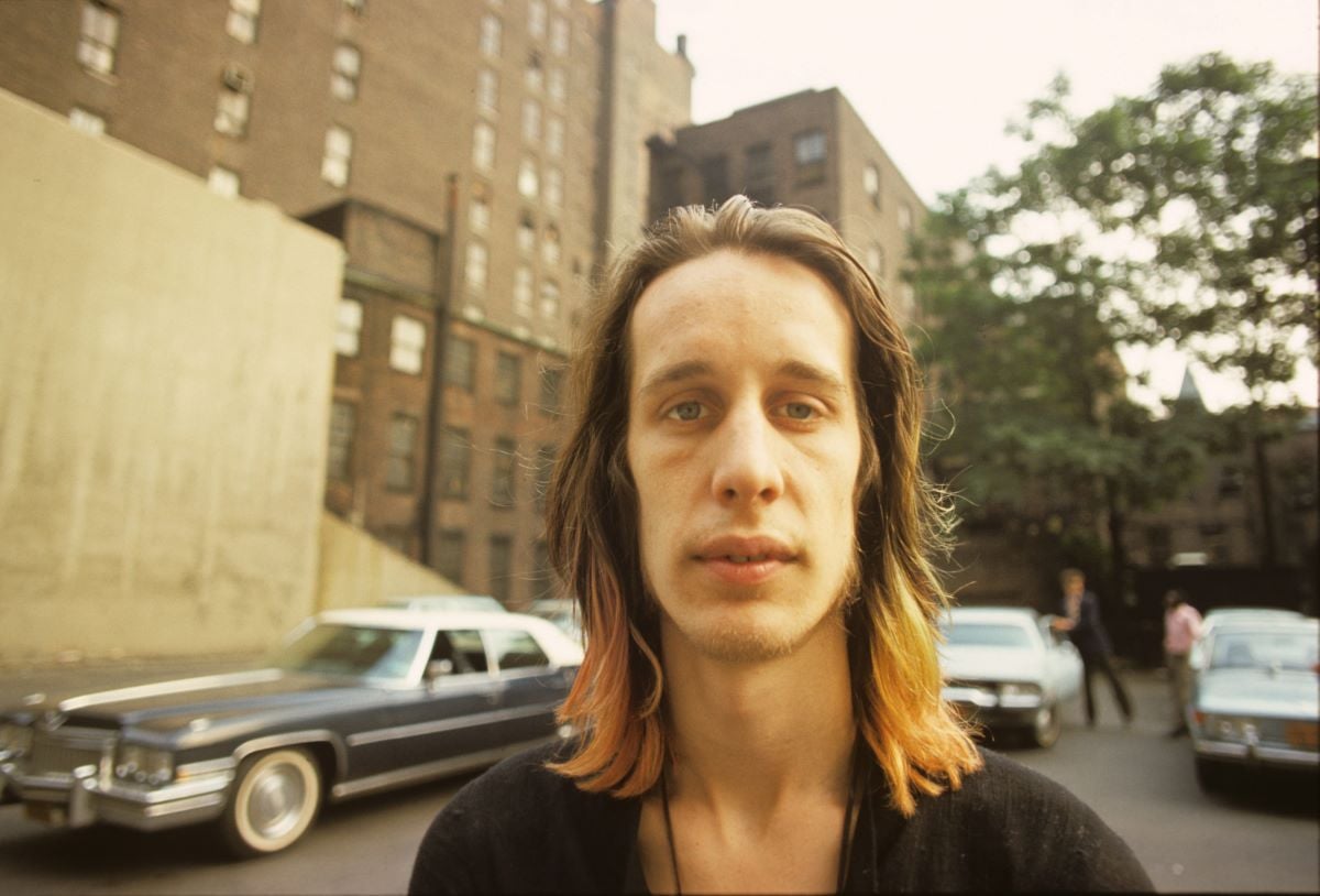 Todd Rundgren stands in front of parked cars and an apartment building.