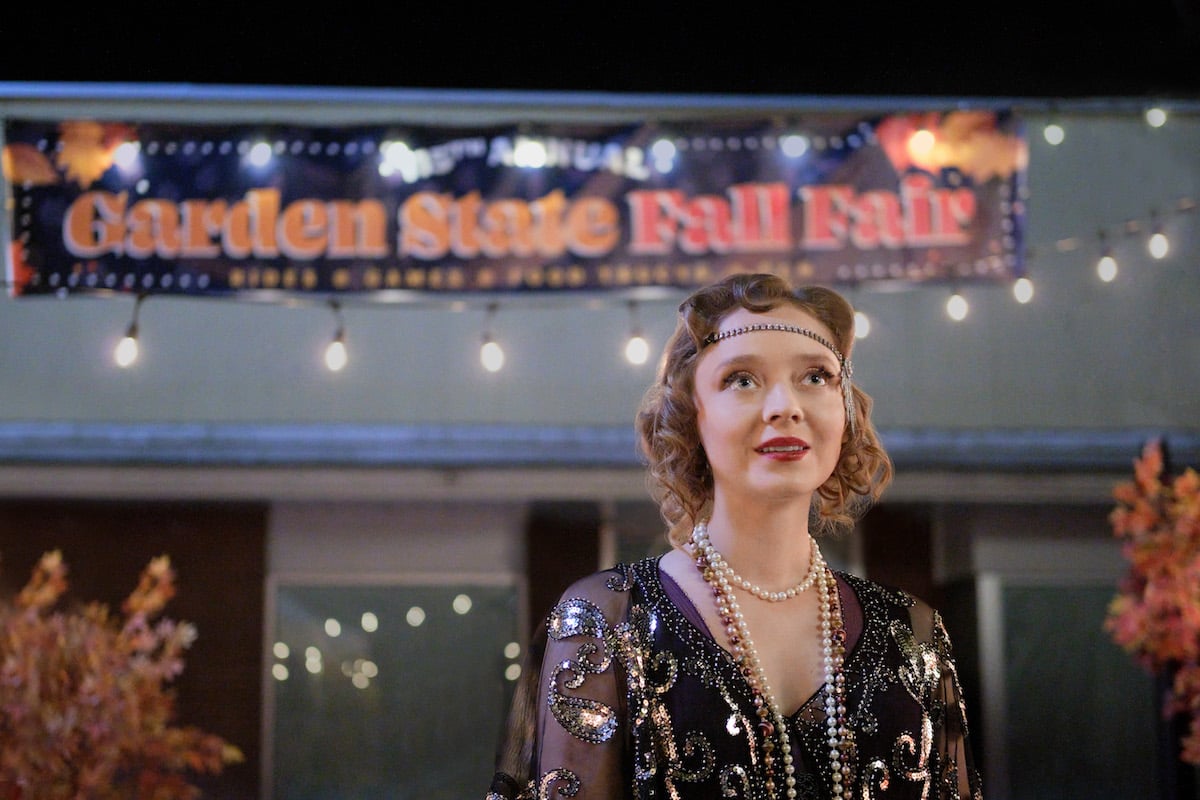 Madeleine Arthur dressed as a 1920s flapper in front of a fall harvest festival sign in Hallmark's '3 Bed, 2 Bath, 1 Ghost'