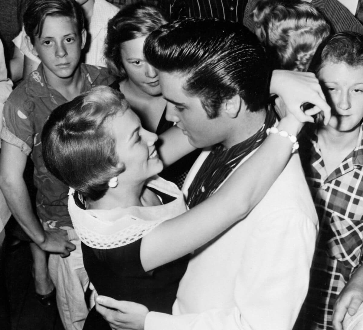 A black and white picture of Anita Wood dancing with her arms around Elvis' neck while a crowd of people watch.