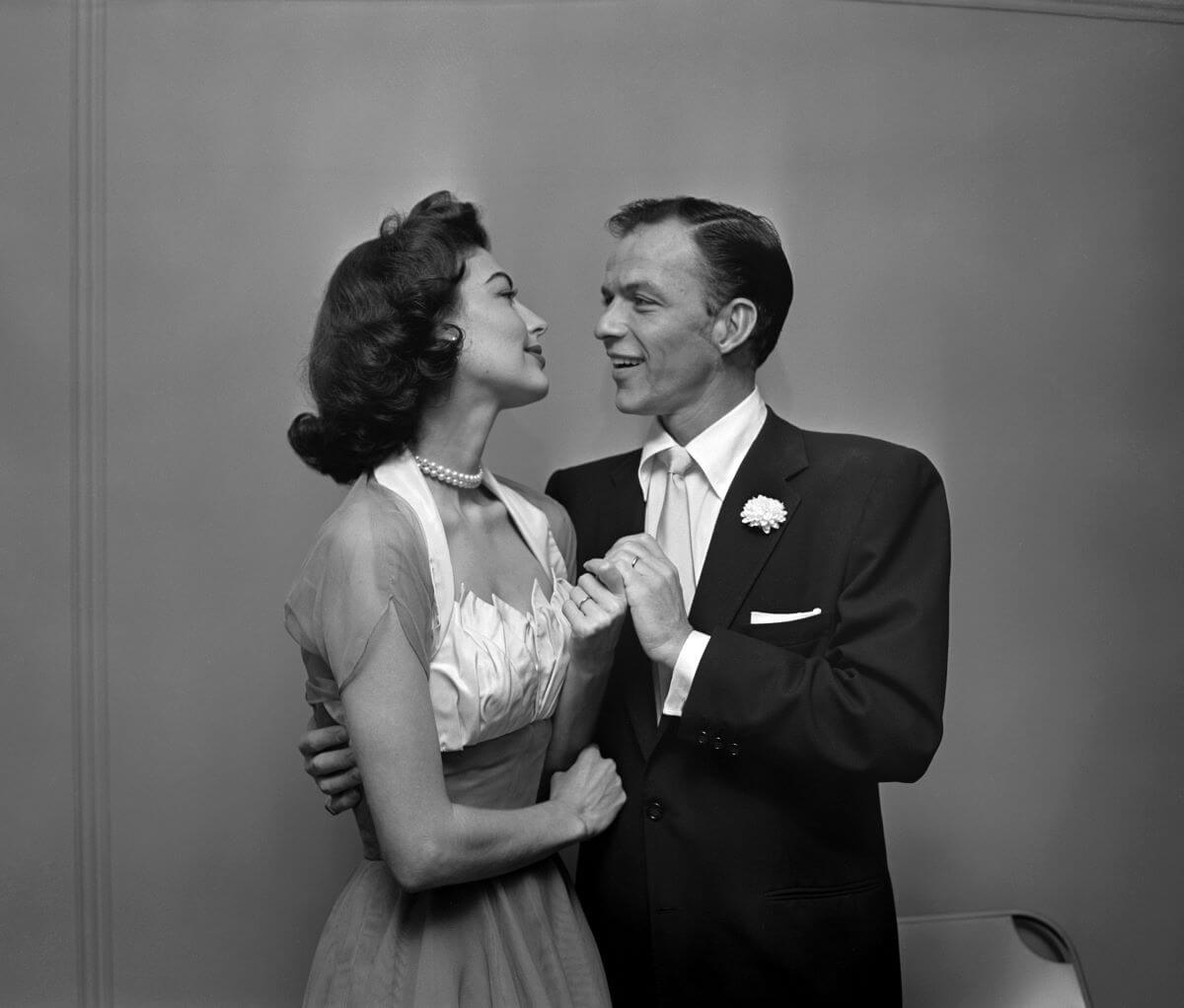 A black and white picture of Ava Gardner and Frank Sinatra holding hands and looking at each other. She wears a dress and he wears a suit.