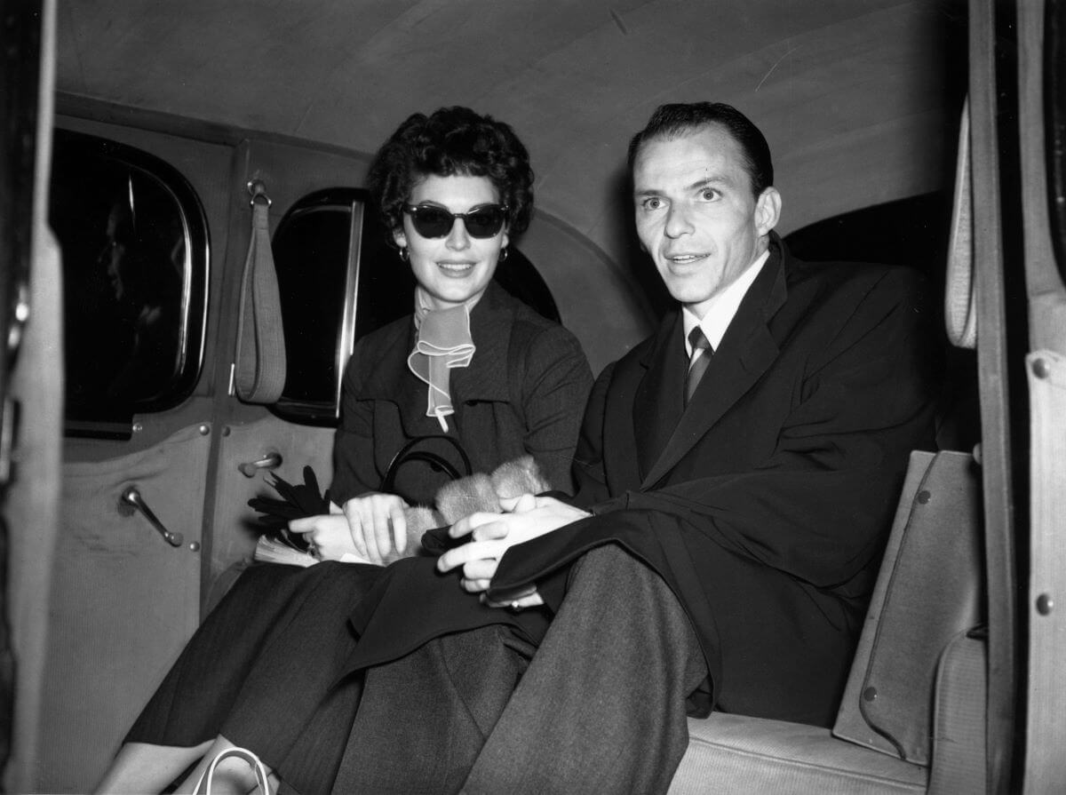 A black and white picture of Ava Gardner and Frank Sinatra sitting in the backseat of a car. She wears sunglasses.