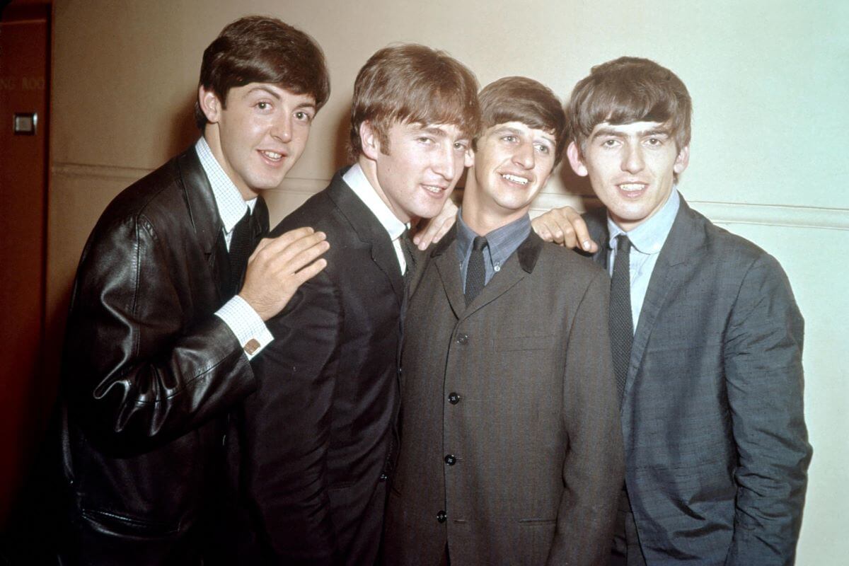 Paul McCartney, John Lennon, Ringo Starr, and George Harrison of The Beatles wearing suits and gathering close together. They lean in and touch each others' shoulders.