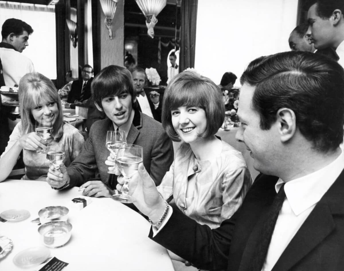 A black and white picture of Pattie Boyd, George Harrison, Cilla Black, and Brian Epstein lifting their glasses at a dinner table. 