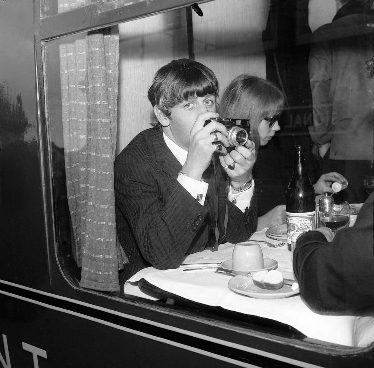 A black and white photo of Ringo Starr sitting at a table on a train and holding a camera. He sits next to Pattie Boyd.