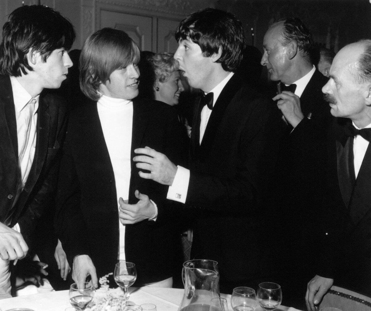 A black and white picture of Keith Richards, Brian Jones, and Paul McCartney talking in a crowded room. They wear tuxedos and stand next to a dinner table.