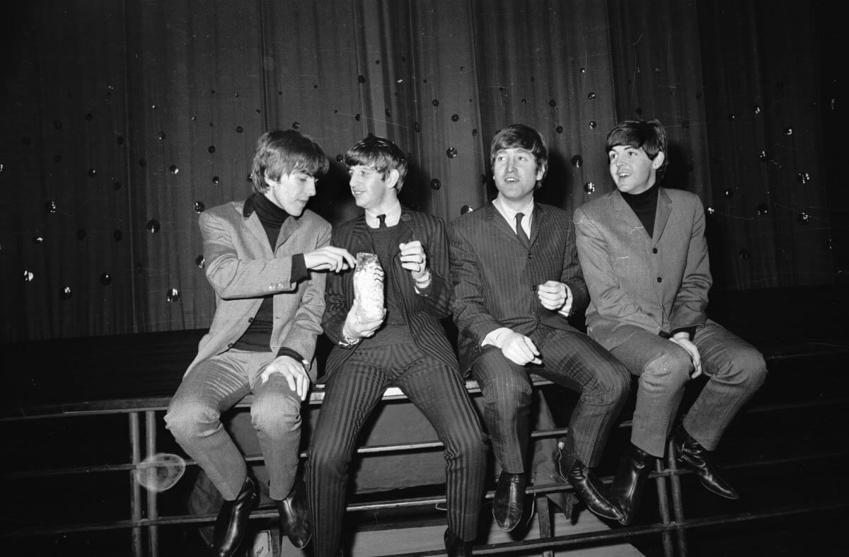 A black and white picture of George Harrison, Ringo Starr, John Lennon, and Paul McCartney sitting on a railing. Starr holds popcorn and Harrison takes some.
