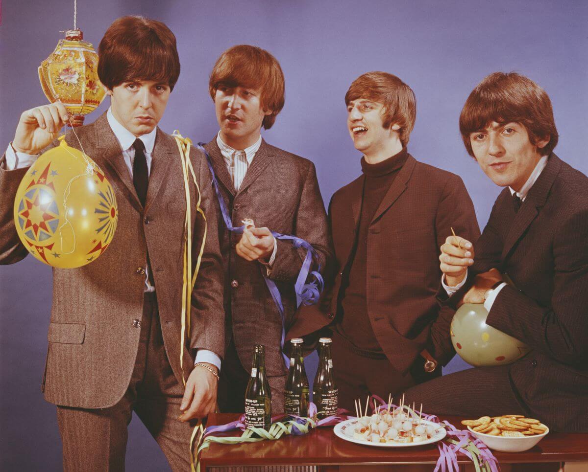 Paul McCartney, John Lennon, Ringo Starr, and George Harrison of The Beatles stand around a table with snacks and drinks. McCartney holds a yellow balloon.