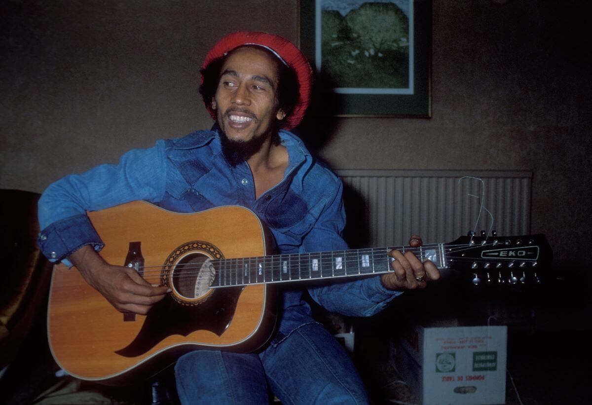 Bob Marley wears a red hat and blue shirt while holding an acoustic guitar.