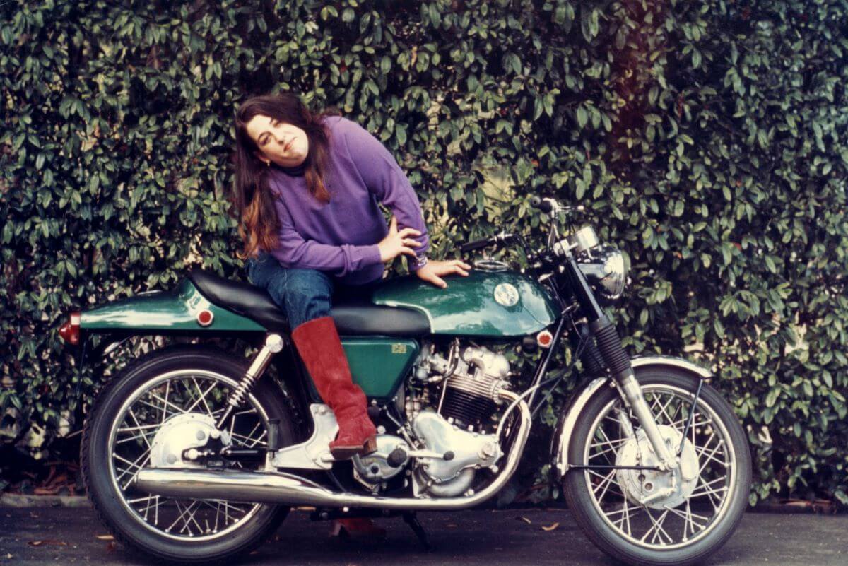 Cass Elliot wears a purple shirt and poses on a green motorcycle.