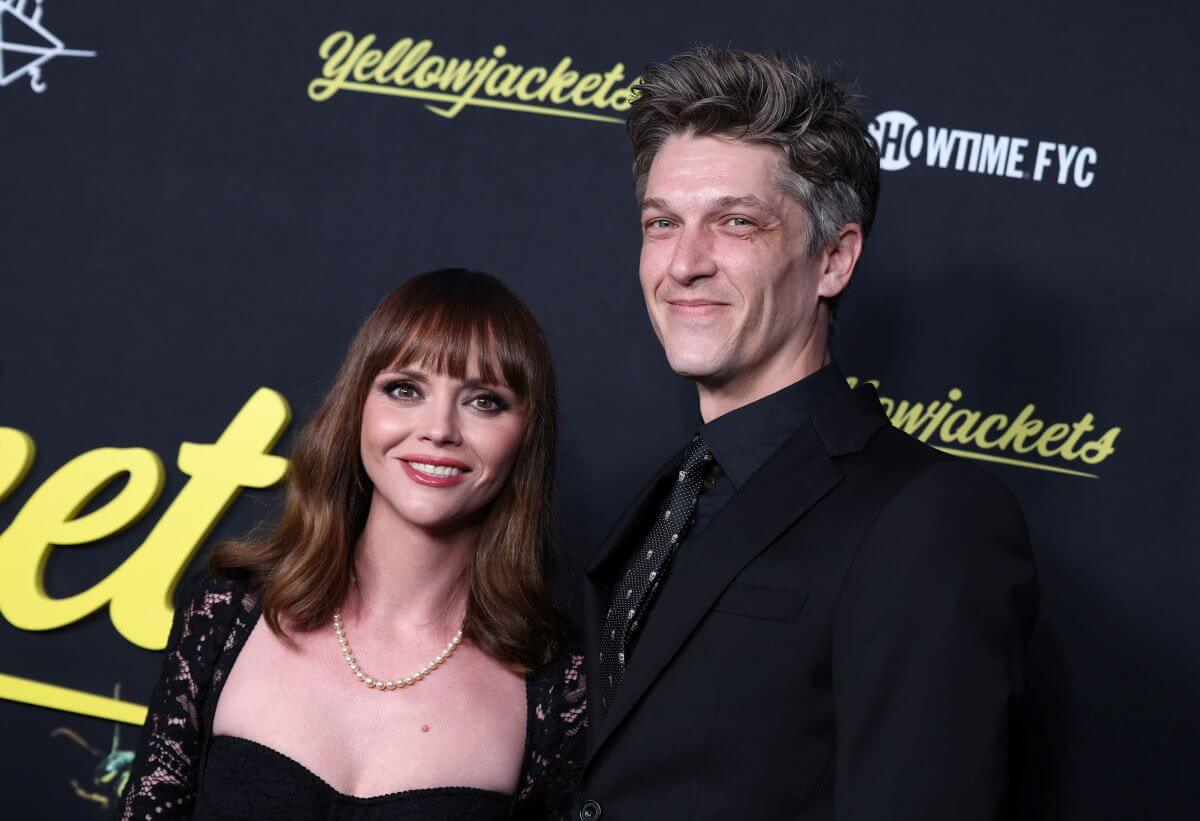Christina Ricci wears a black dress and a pearl necklace. She stands next to Mark Hampton who wears a black suit and tie. They stand in front of signs for 'Yellowjackets.'