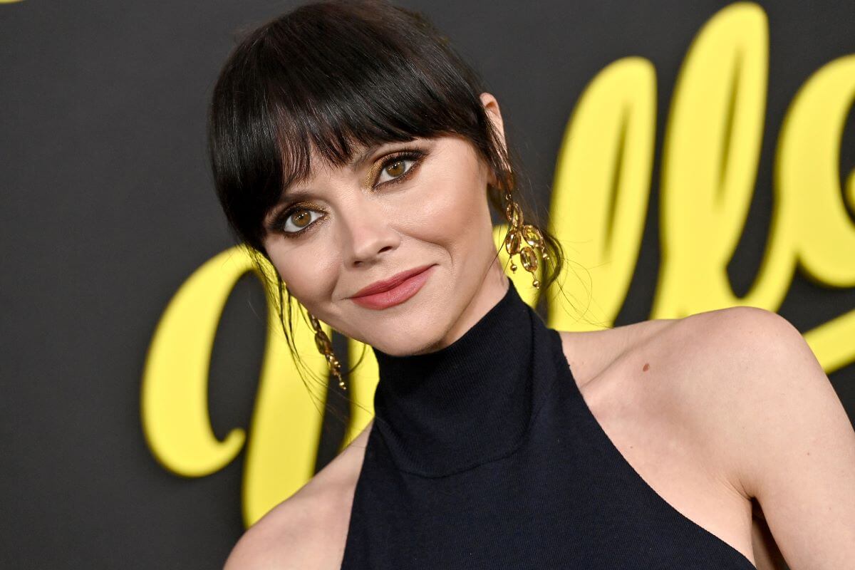 Christina Ricci wears a black high-necked dress and dangly earrings. She stands in front of a poster for 'Yellowjackets.'