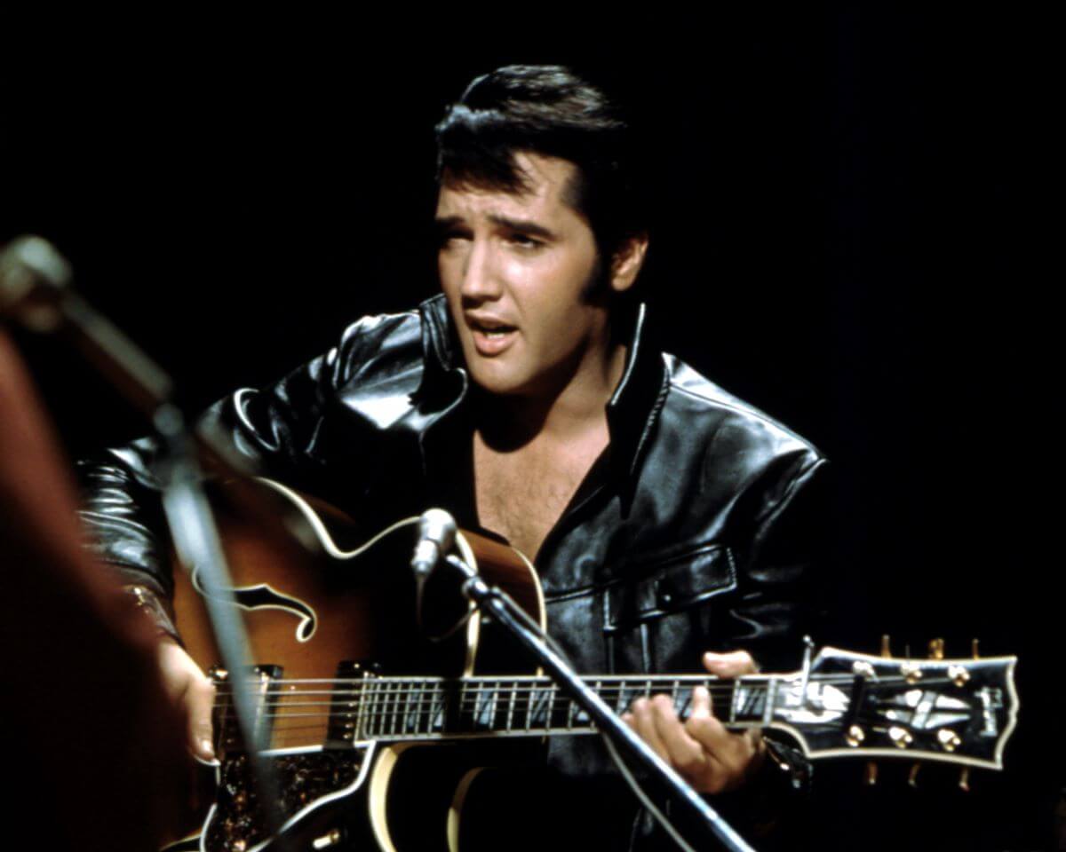 Elvis Presley wears a leather jacket and sits while strumming an acoustic guitar.