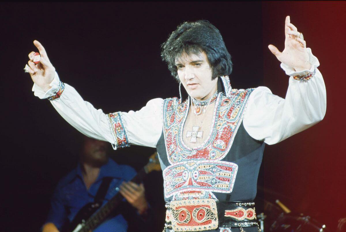 Elvis Presley stands on a stage and holds up his arms.