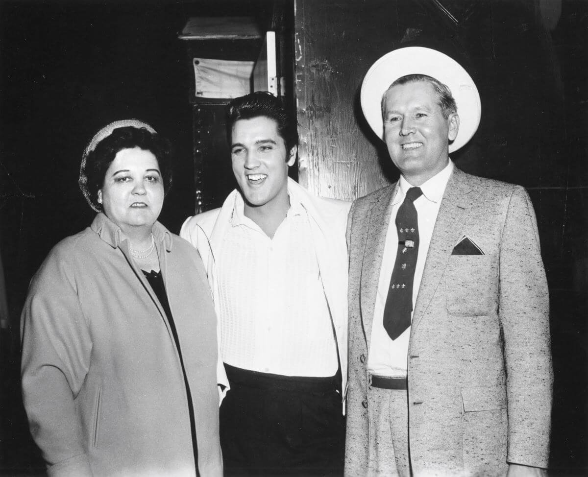A black and white picture of Gladys, Elvis, and Vernon Presley standing together. Elvis has his arms around his parents.