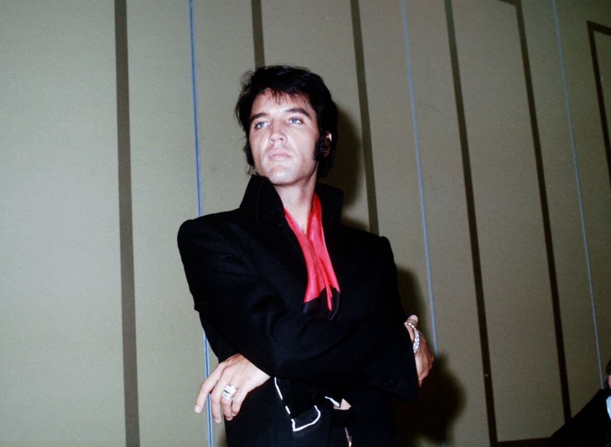 Elvis Presley wears a black jacket with a red tie and stands with his arms crossed.