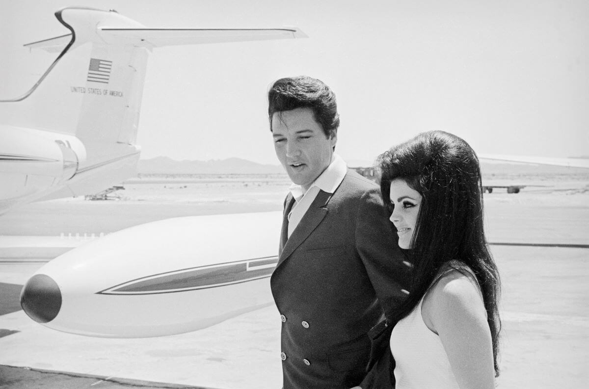 A black and white picture of Elvis and Priscilla Presley standing on a tarmac in front of an airplane.