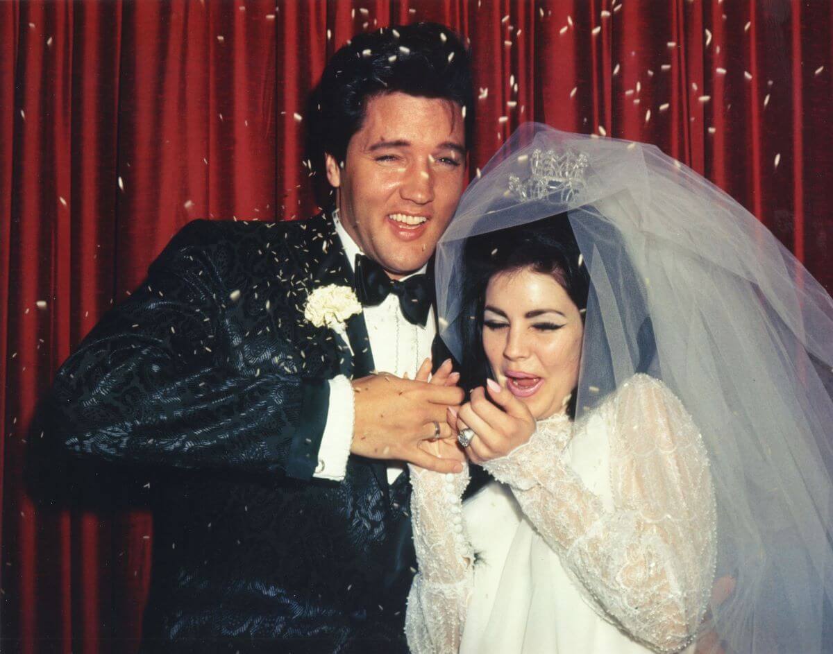 Elvis and Priscilla Presley stand in front of a red curtain and brace themselves against confetti. He wears a tuxedo and she wears her wedding dress and veil.