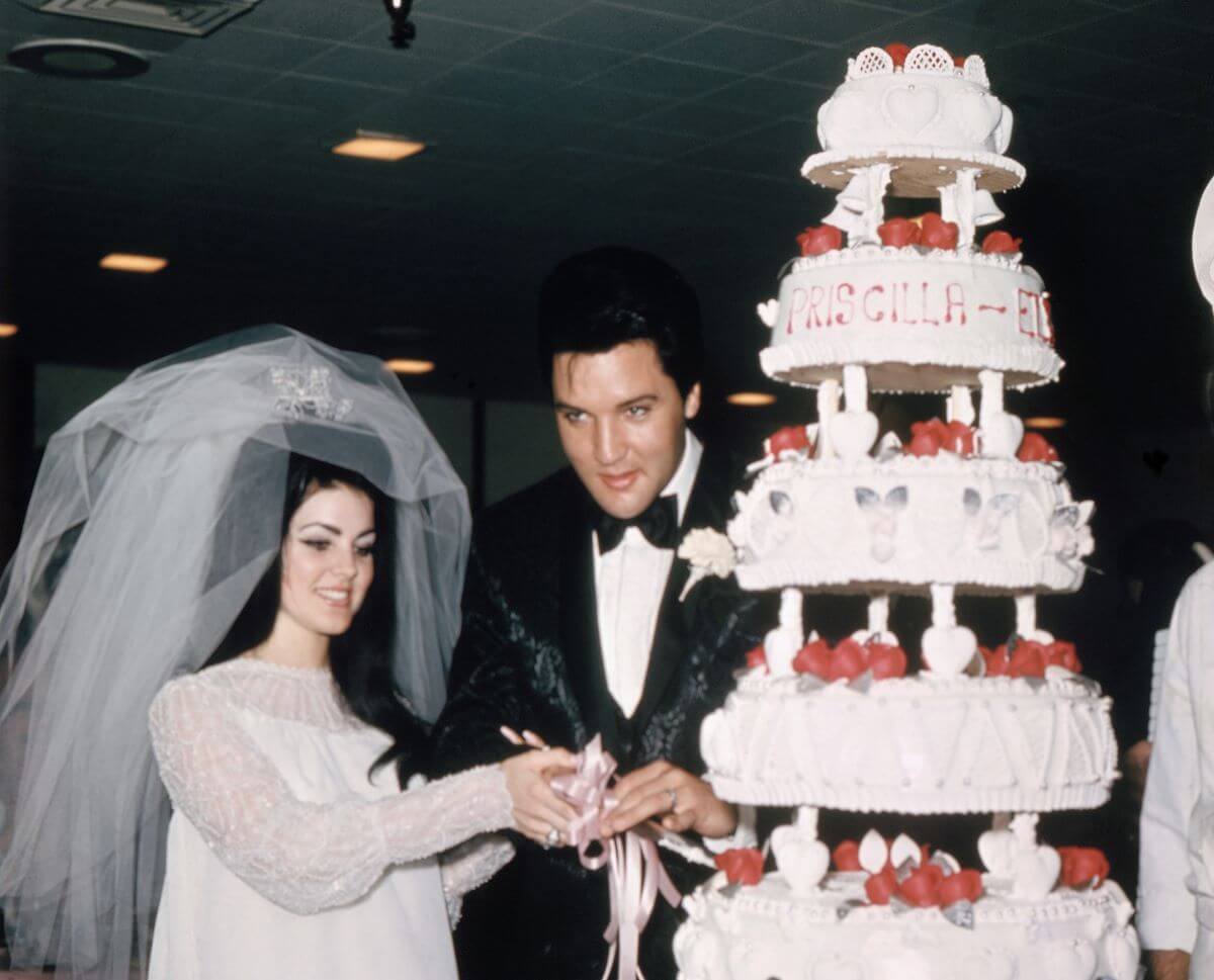 Priscilla and Elvis Presley cut into their wedding cake. She wears her wedding dress and veil and he wears a tuxedo.