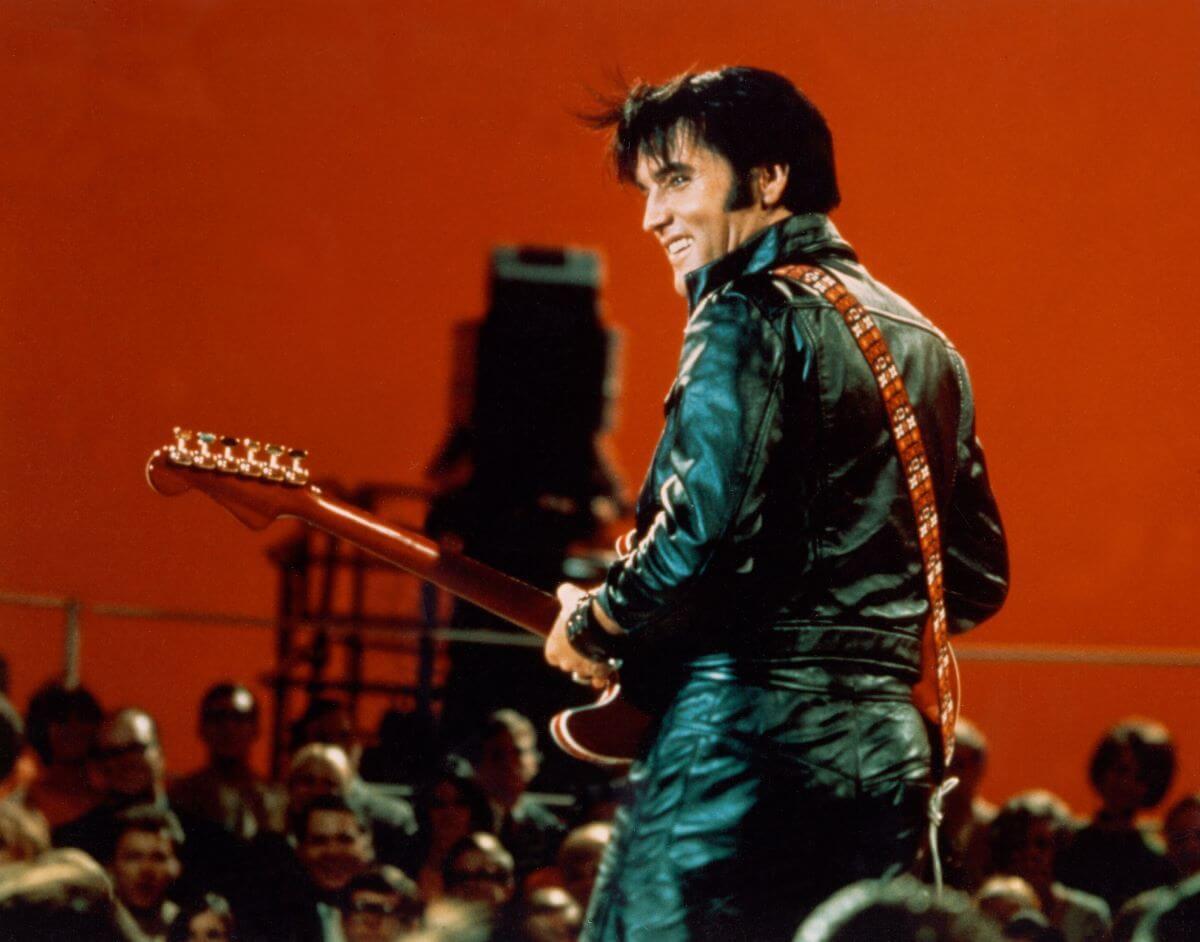 Elvis wears a black leather jacket and holds a guitar. He stands on a stage in a room with red walls.