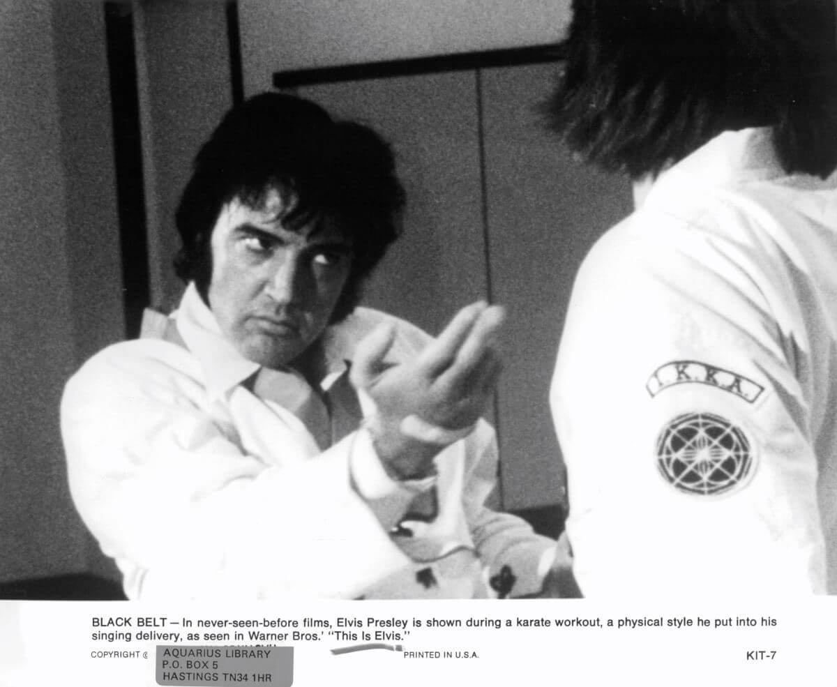 A black and white picture of Elvis holding his hand up in a karate workout. A description at the bottom of the image notes that the image is from the film 'This Is Elvis.'