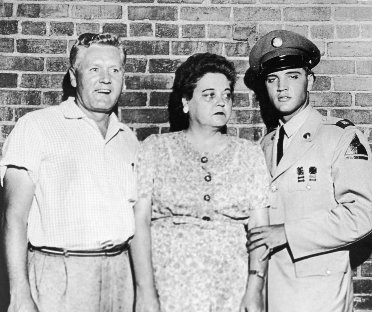 A black and white picture of Vernon, Gladys, and Elvis Presley standing in front of a brick wall. Elvis wears his Army uniform.