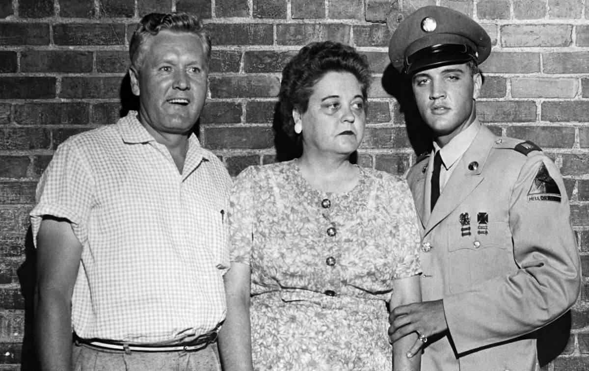 A black and white picture of Vernon, Gladys, and Elvis Presley standing in front of a brick wall. Elvis wears his Army uniform.