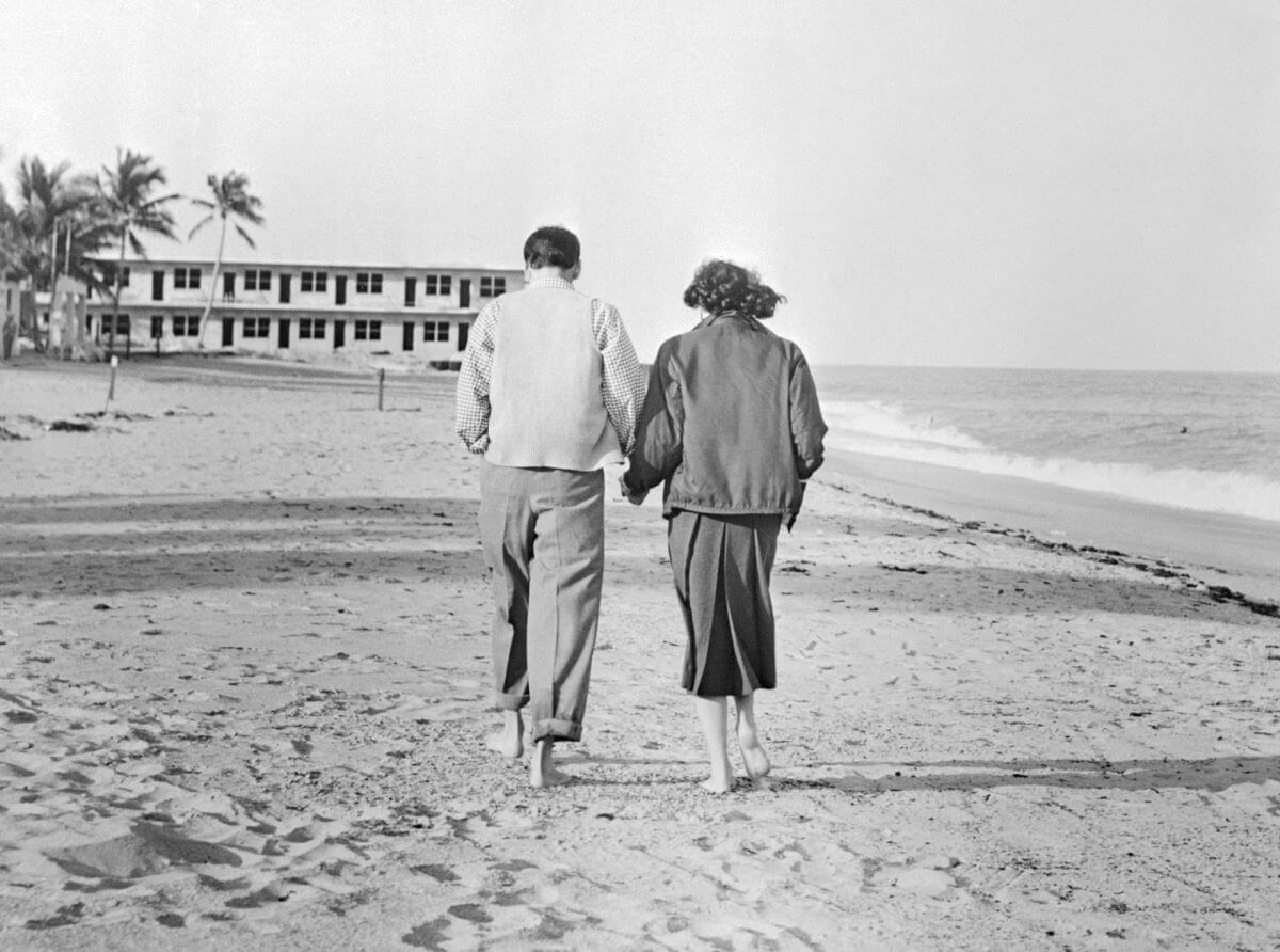 A black and white picture of Frank Sinatra and Ava Gardner walking barefoot on a beach. The camera captures their backs.