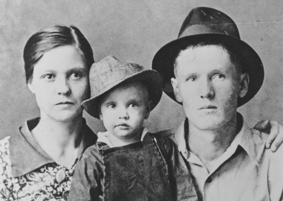 A black and white picture of Gladys and Vernon Presley posing with a toddler Elvis between them. Elvis and Vernon wear hats.