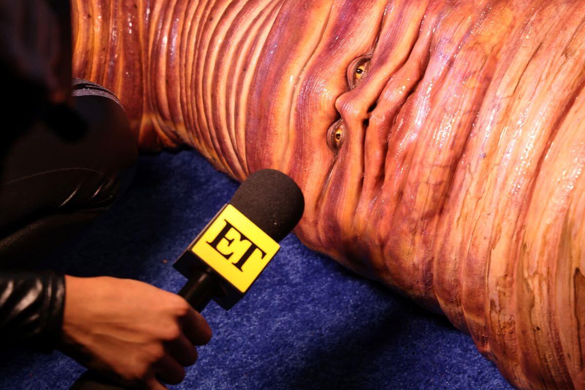 Heidi Klum lays on the ground in a worm costume. A reporter holds an ET microphone up to her.