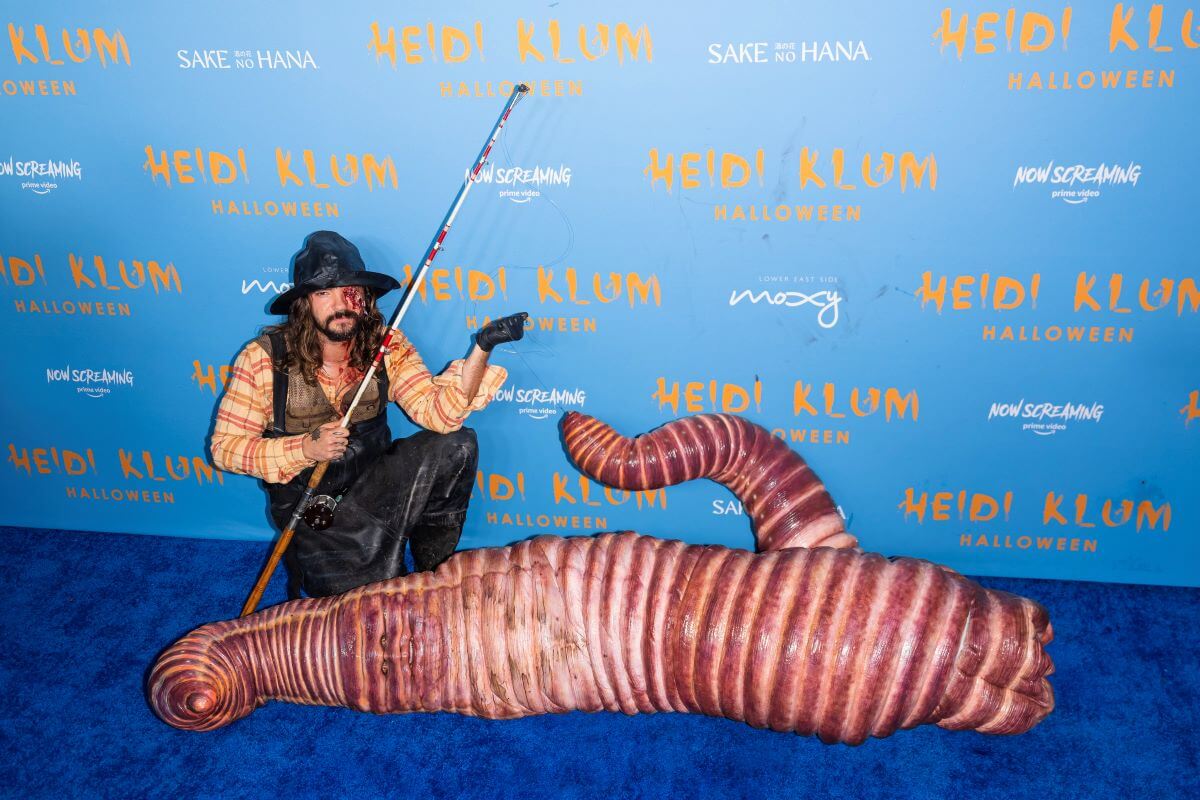 Heidi Klum lays on the ground in a worm costume while a man holds a fishing pole next to her.