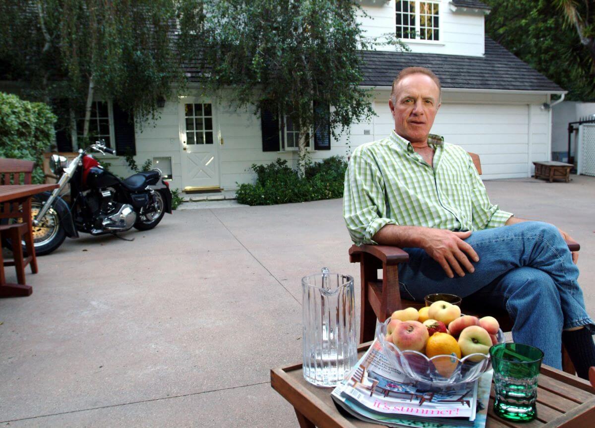James Caan wears a green shirt and sits in his driveway in front of his house and a motorcycle. A table with a bowl of apples is in front of him.