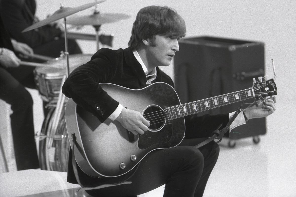 A black and white picture of John Lennon wearing a suit and sitting on a stage while strumming an acoustic guitar.