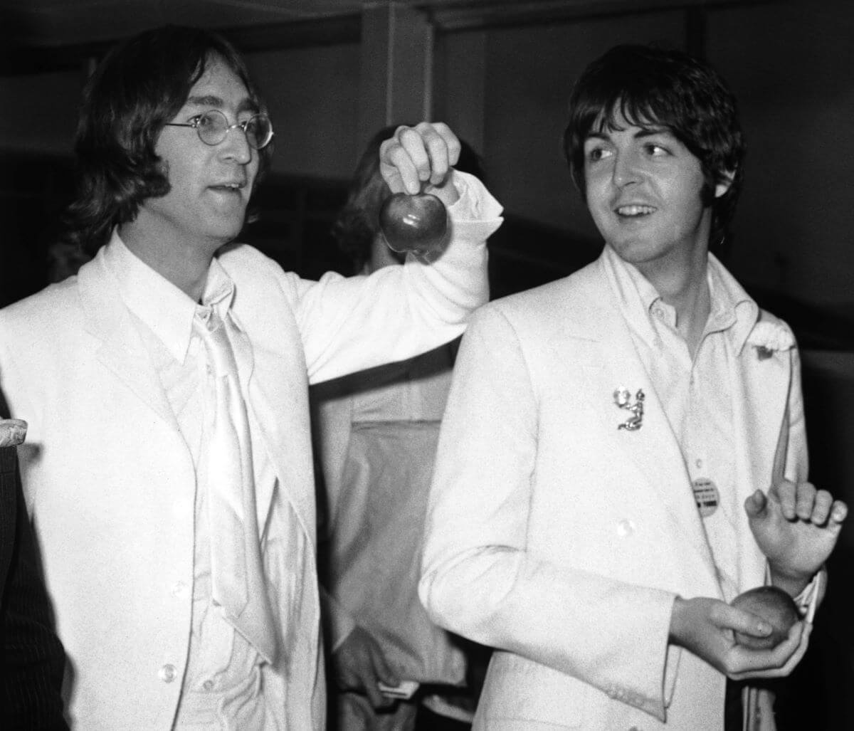 A black and white picture of John Lennon and Paul McCartney wearing white suits and holding apples.