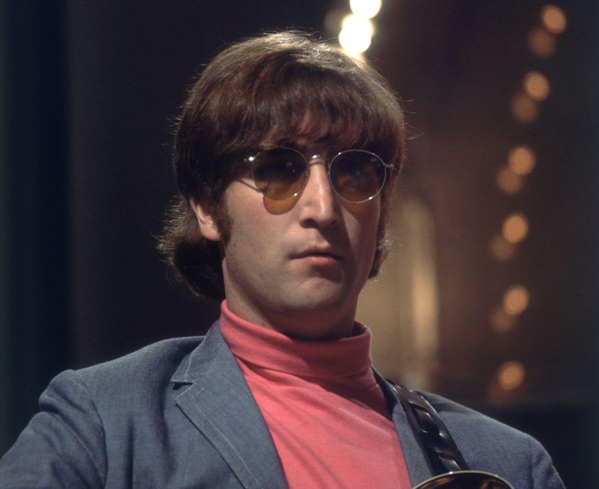 John Lennon wears a turtleneck, blue jacket, and sunglasses.