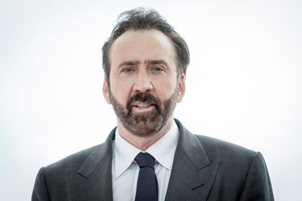 Nicolas Cage wears a suit and tie and stands in front of a blank white background.
