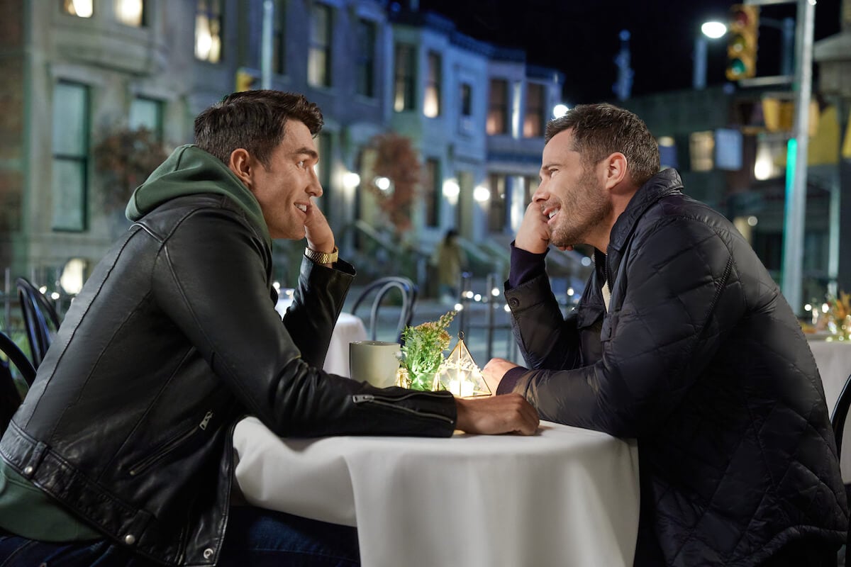 Luke Macfarlane and Peter Porte holding hands and gazing at each other across a table in the Hallmark movie 'Notes of Autumn'