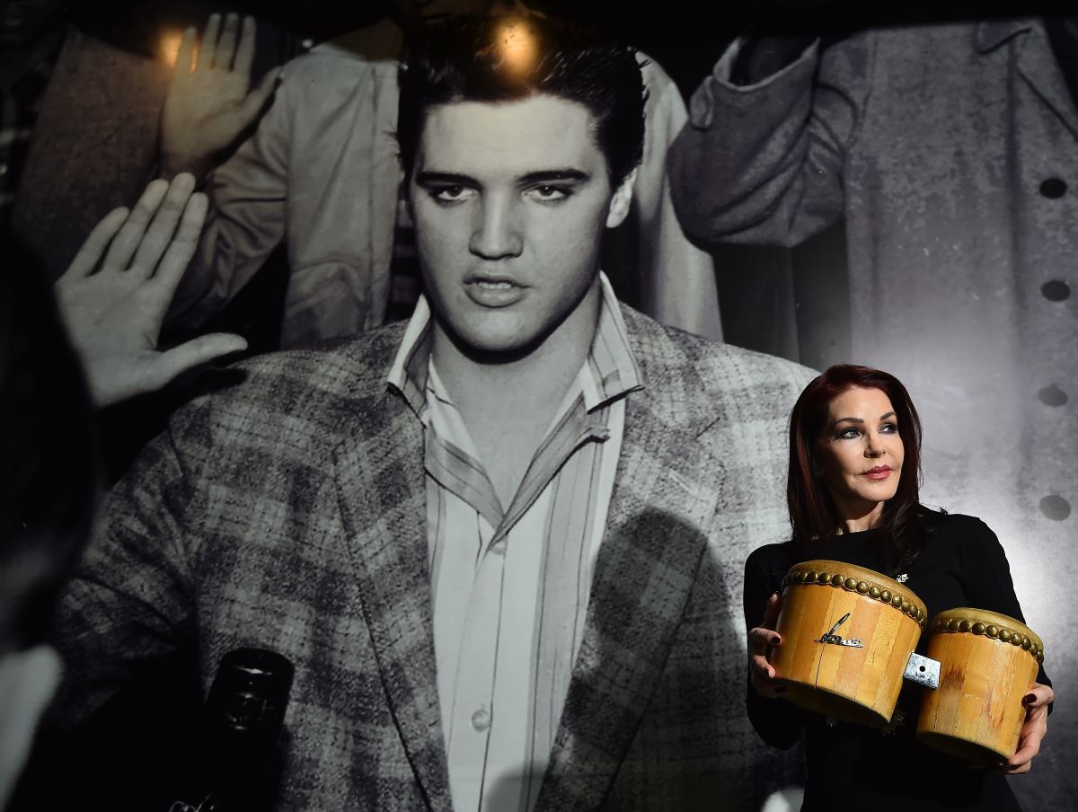 Priscilla Presley holds a pair of bongos and stands in front of a large black and white photo of Elvis Presley.