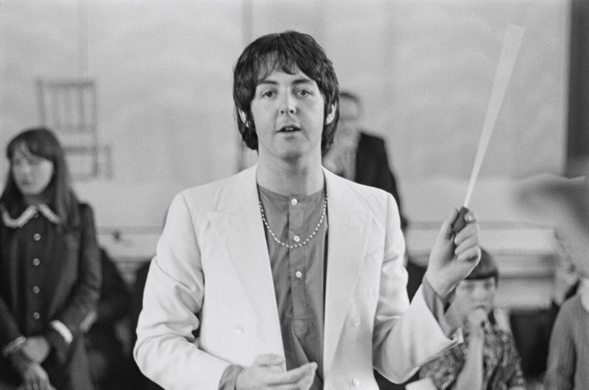 A black and white picture of Paul McCartney wavong a conductor's baton and standing in front of several people.