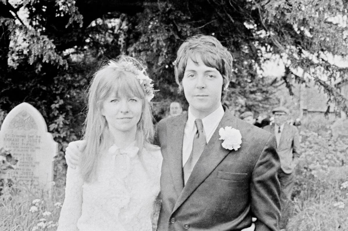 A black and white picture of Paul McCartney standing with his arm around Jane Asher's shoulders. They stand outside a church. There is a gravestone behind them. 