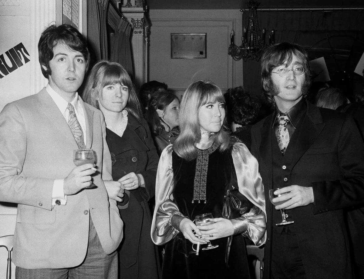 A black and white picture of Paul McCartney, Jane Asher, Cynthia Lennon, and John Lennon. They stand together at a party and hold drinks.