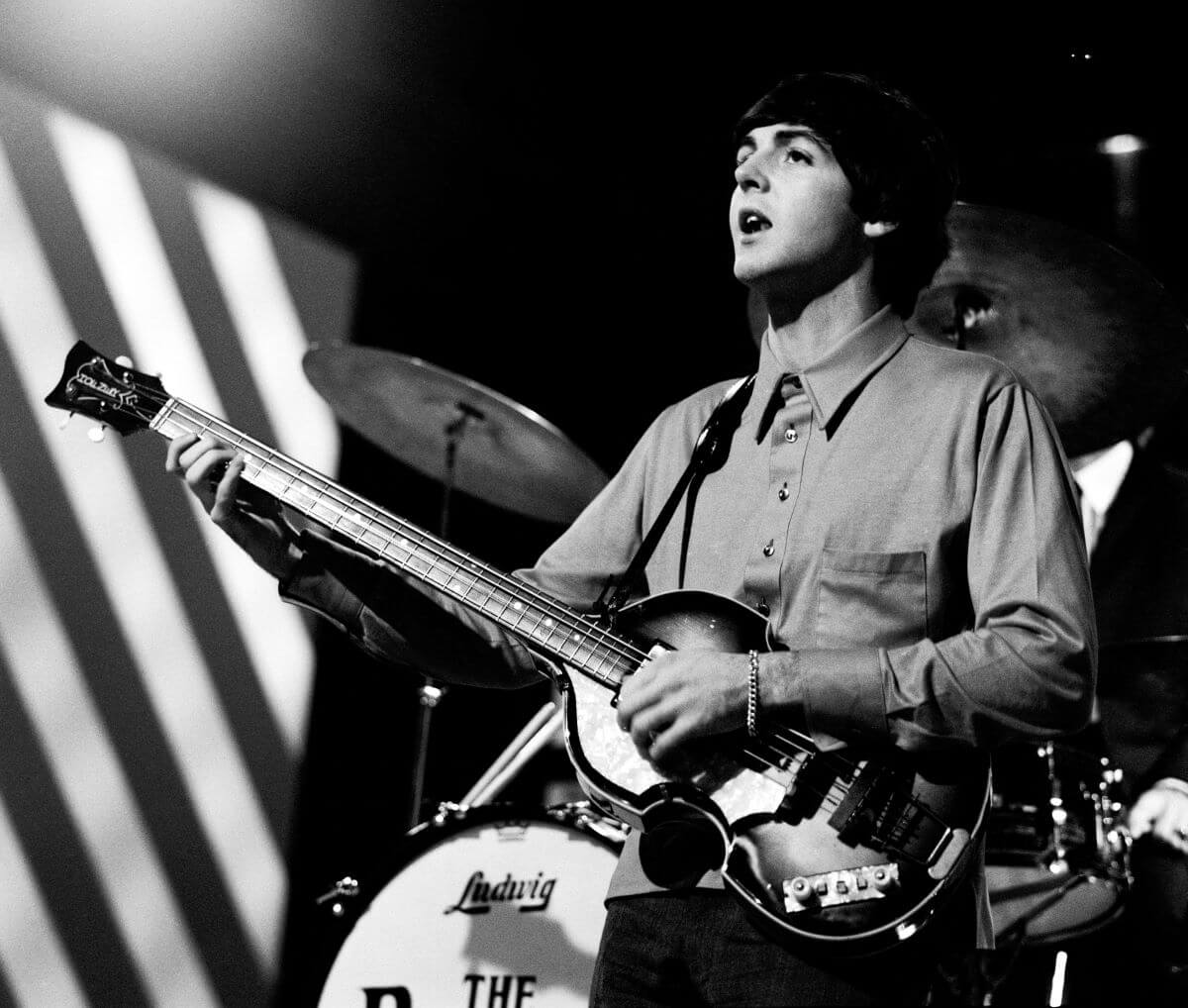 A black and white picture of Paul McCartney standing in front of a drum set and holding a guitar.