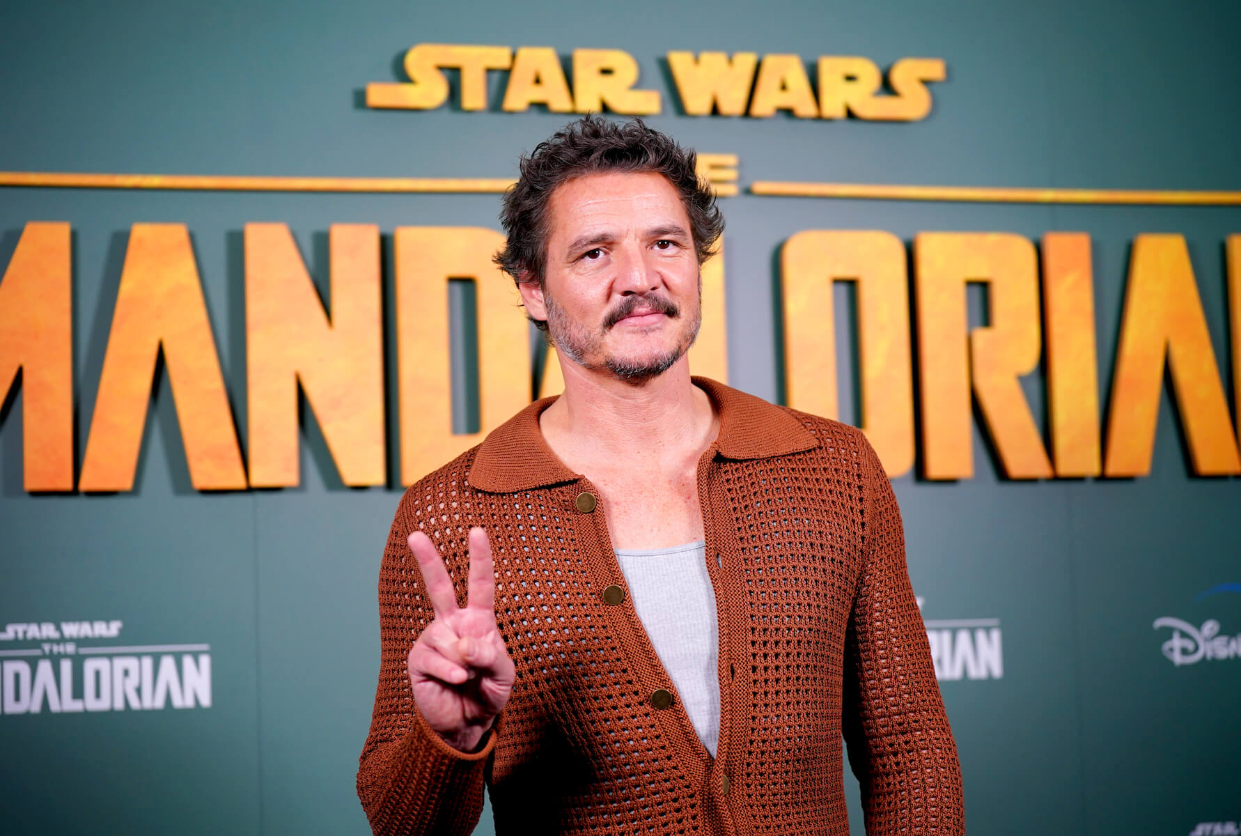 Pedro Pascal holding his hand up in a peace sign in front of 'The Mandalorian' logo