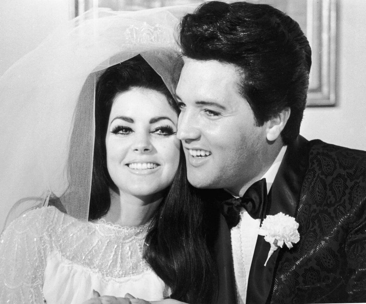 A black and white picture of Priscilla Presley wearing her veil and wedding dress and Elvis Presley wearing a tuxedo.
