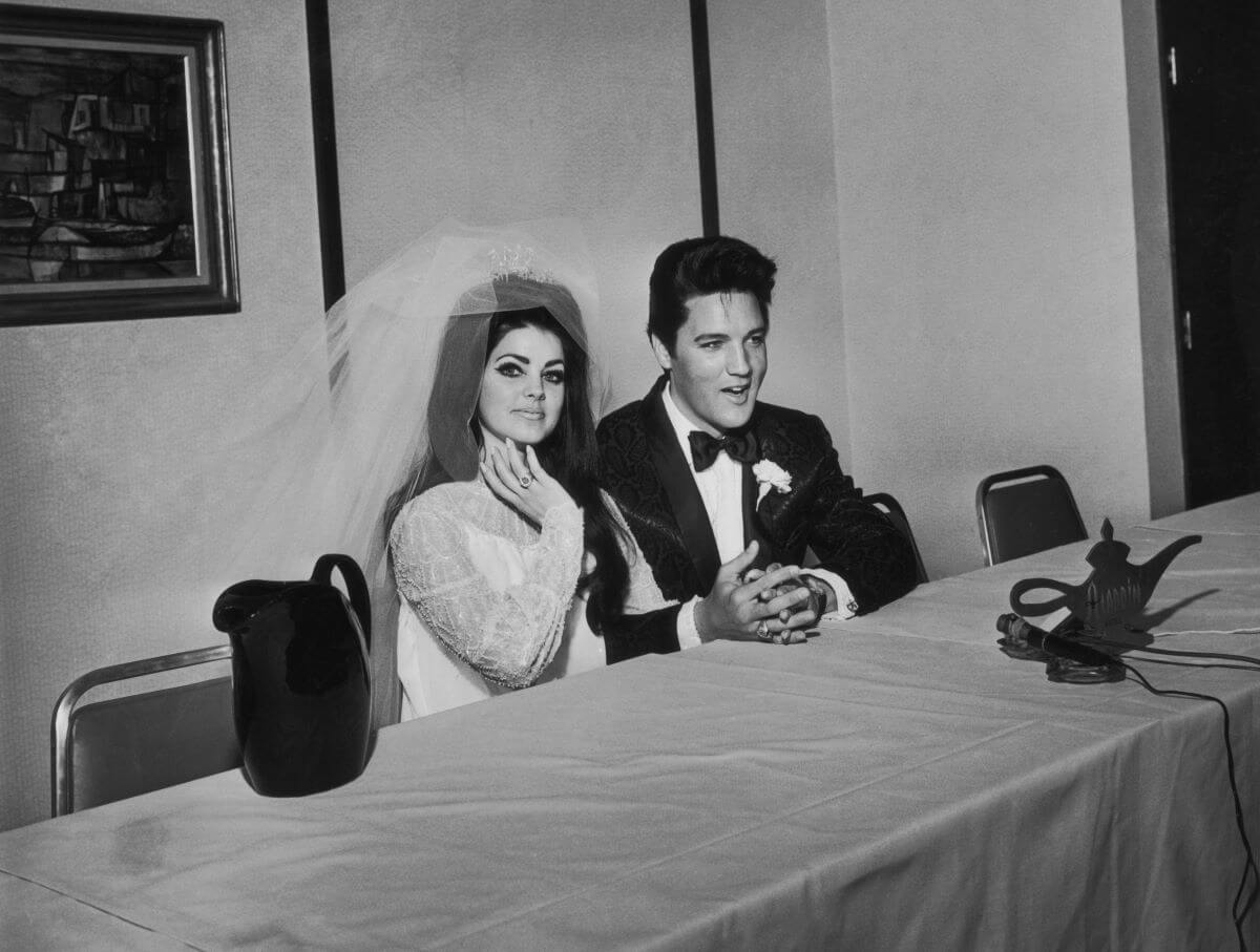 A black and white picture of Priscilla and Elvis Presley sitting on the same side of a long table on their wedding day. She wears her dress and veil and he wears a tuxedo.