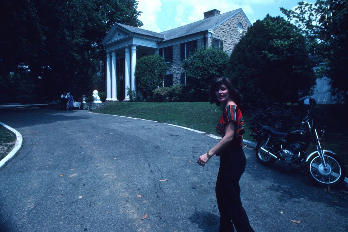 Priscilla Presley walks toward Graceland. She stands near a motorcycle.