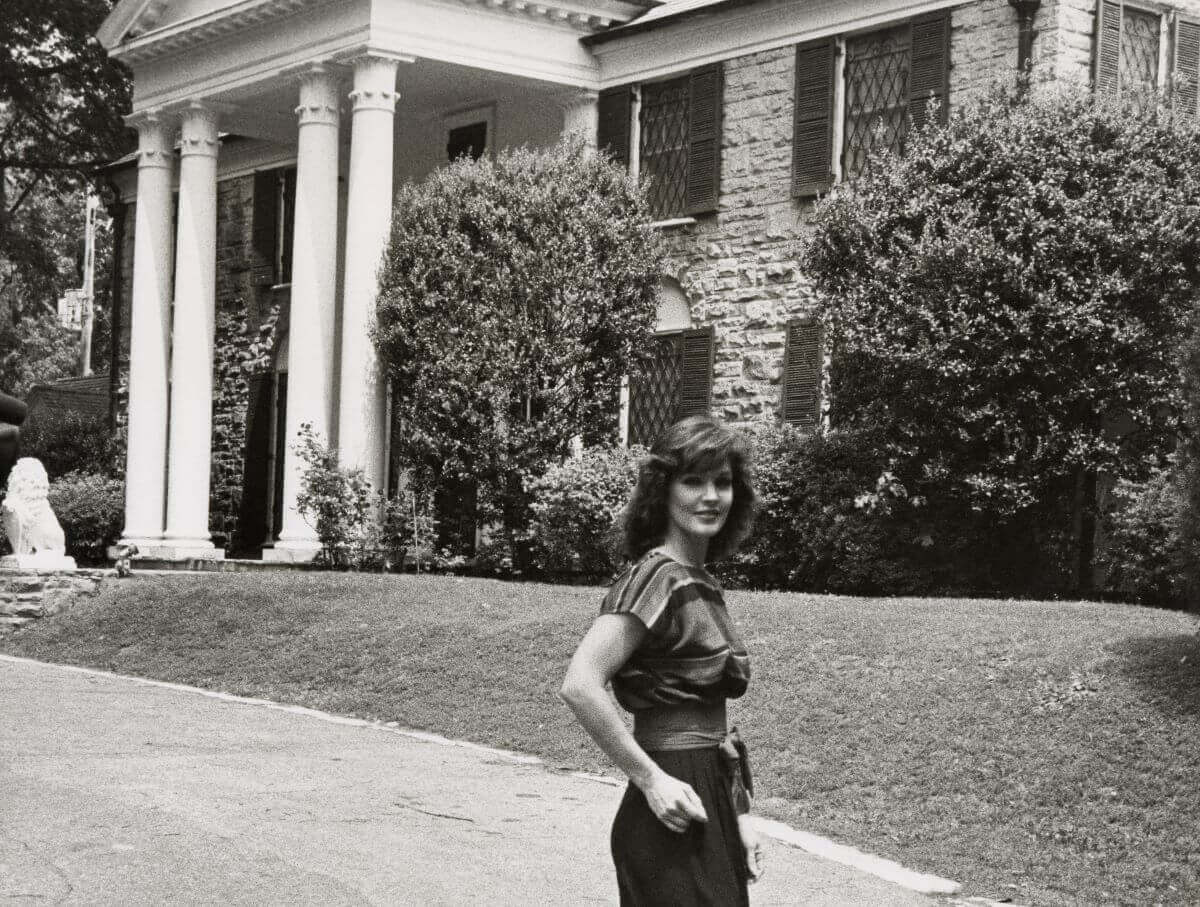A black and white picture of Priscilla Presley standing with her hands in her pockets in front of Graceland.