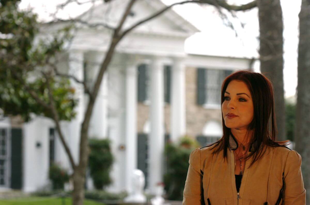 Priscilla Presley wears a tan jacket and stands in front of Graceland.