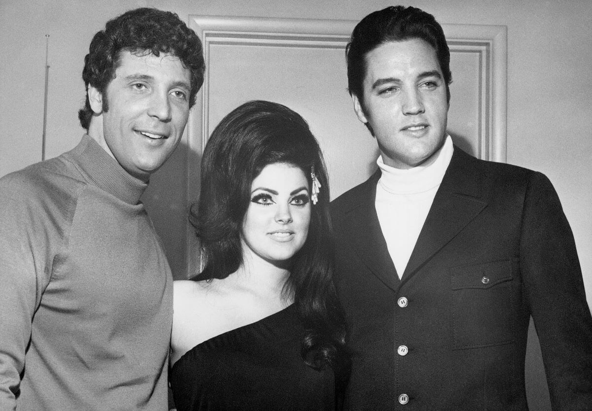 A black and white picture of Priscilla Presley standing in between Tom Jones and Elvis Presley. She wears a one-shouldered dress and they both wear turtlenecks. 