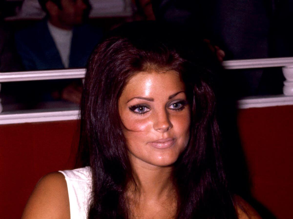 Priscilla Presley sits in a booth and wears a white shirt.