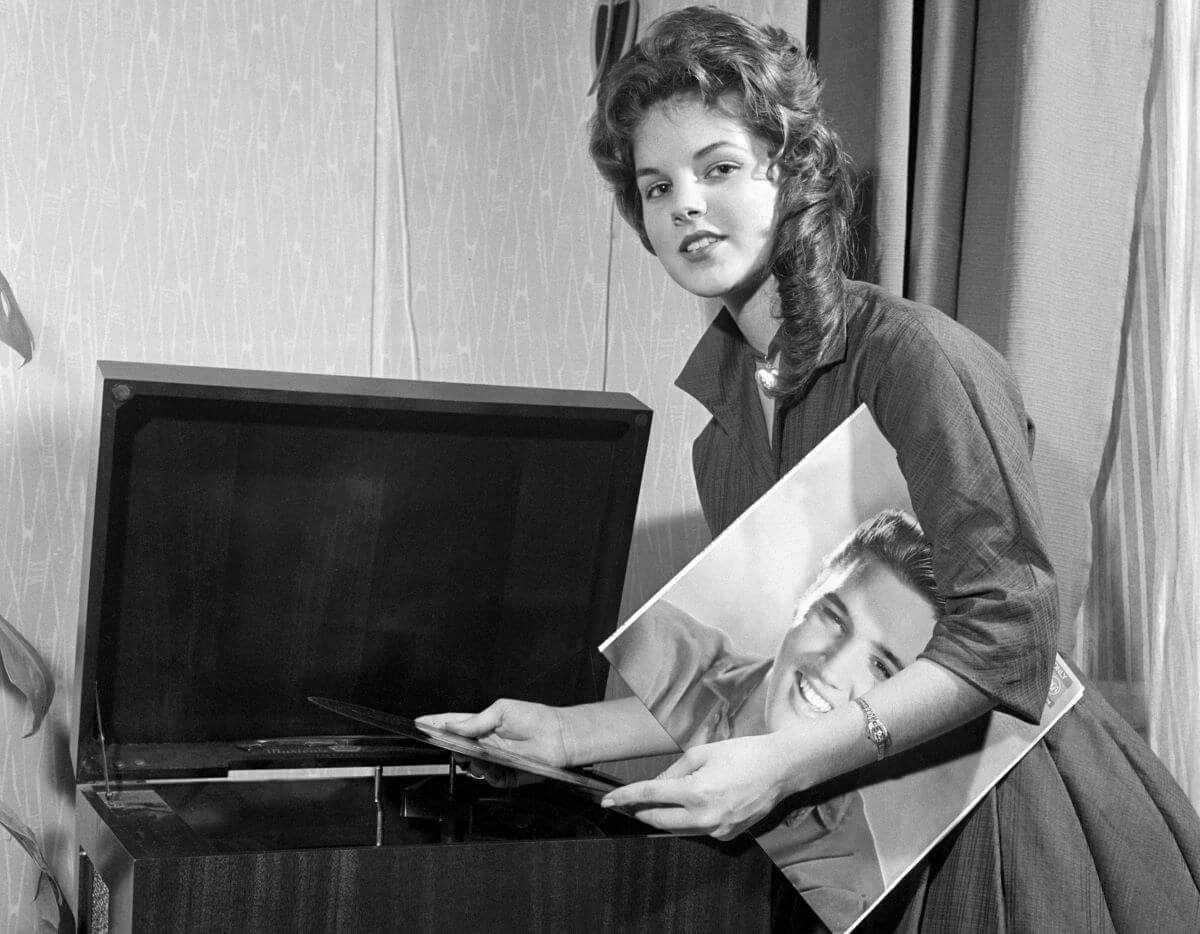 A black and white picture of Priscilla Presley holding an Elvis record in front of a record player.